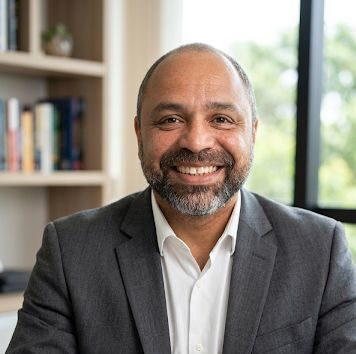 Ari Lima Ari lima at a table wearing a grey suit , window and shelving with books in the background